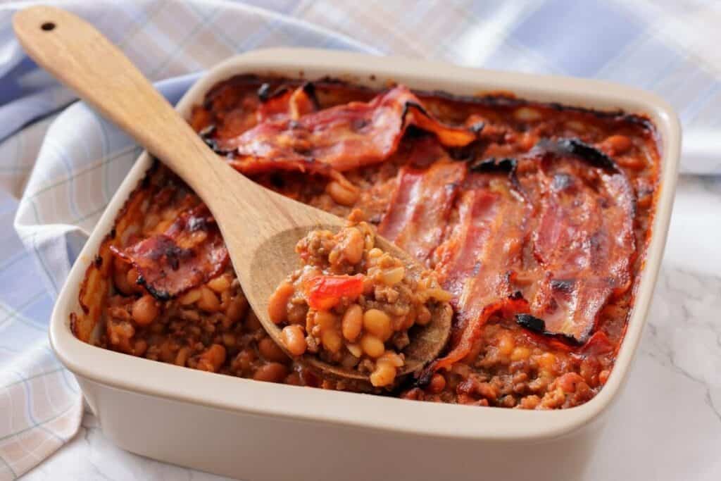 A square baking dish filled with baked beans, ground meat, and topped with strips of cooked bacon; a wooden spoon holds a serving.