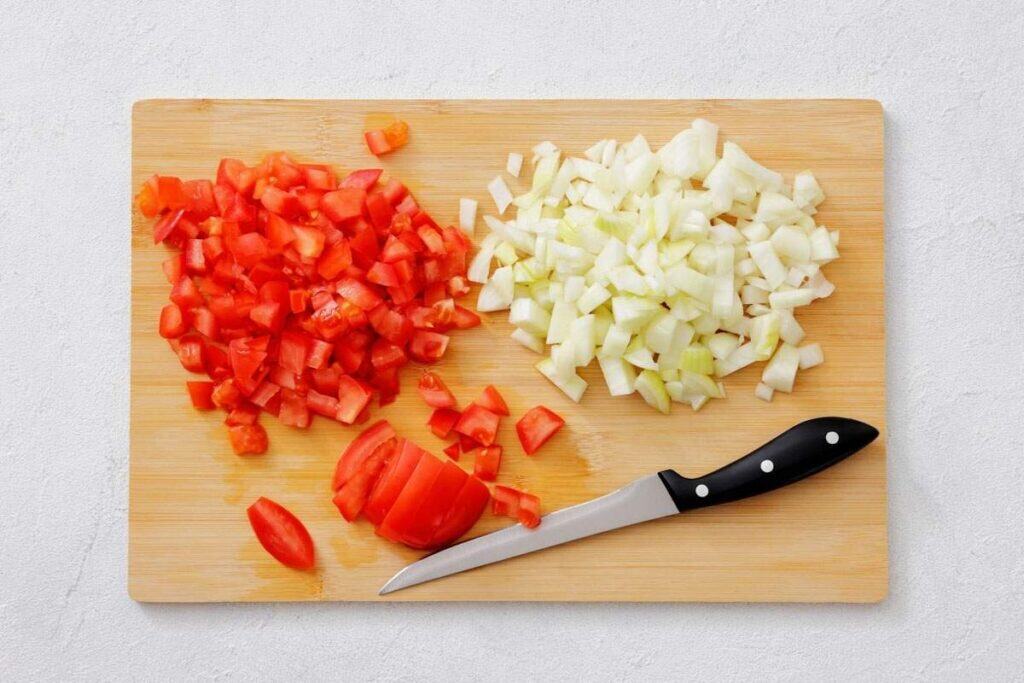 A cutting board with chopped tomatoes, chopped onions, a partially sliced tomato, and a kitchen knife.