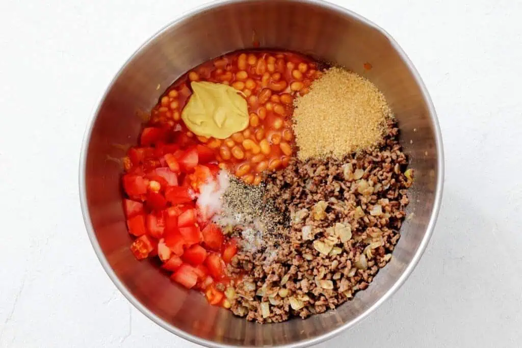 A metal bowl containing baked beans, diced tomatoes, ground beef with onions, brown sugar, mustard, salt, and pepper, all separated in sections.