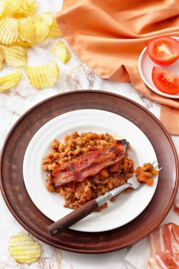 A white plate with baked beans and a strip of bacon sits on a wooden charger. Nearby are potato chips, sliced tomatoes, and an orange napkin.