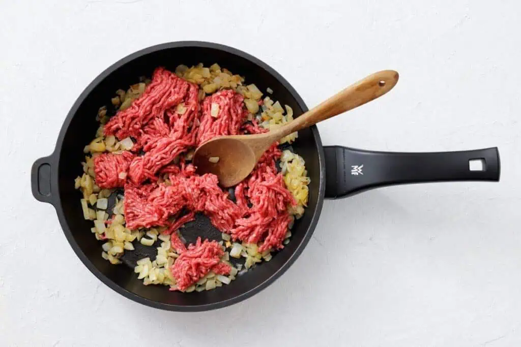 Ground beef and chopped onions being cooked in a black frying pan with a wooden spoon on a white surface.