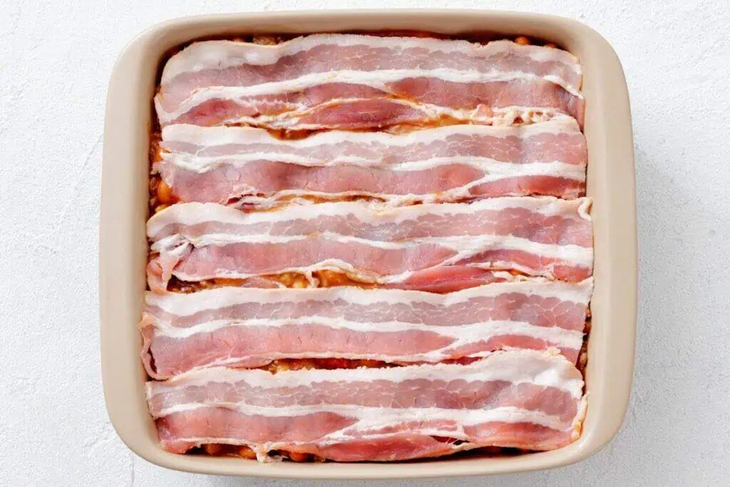 A square baking dish filled with uncooked bacon strips arranged in a single layer, covering the surface.
