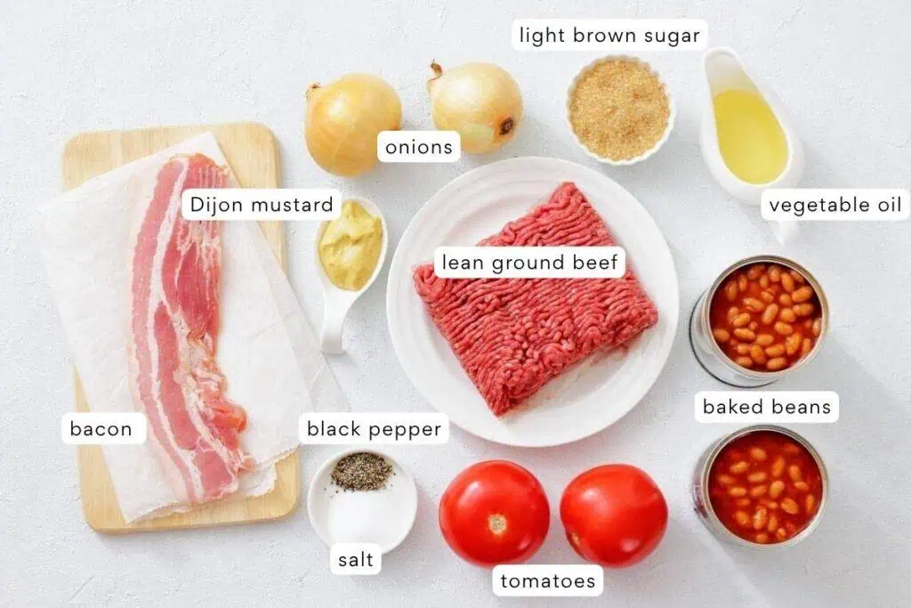 Ingredients for a recipe laid out on a white surface: bacon, Dijon mustard, onions, light brown sugar, vegetable oil, lean ground beef, black pepper, salt, tomatoes, and baked beans.