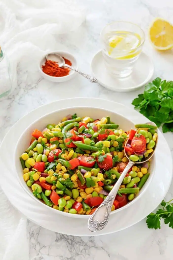 A bowl of mixed vegetable salad with green beans, corn, edamame, and red bell peppers, garnished with herbs, next to a glass of water with lemon and a dish of red spice.