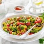 A white bowl filled with a colorful vegetable salad, including edamame, corn, cherry tomatoes, green beans, and red peppers, with a spoon resting inside.