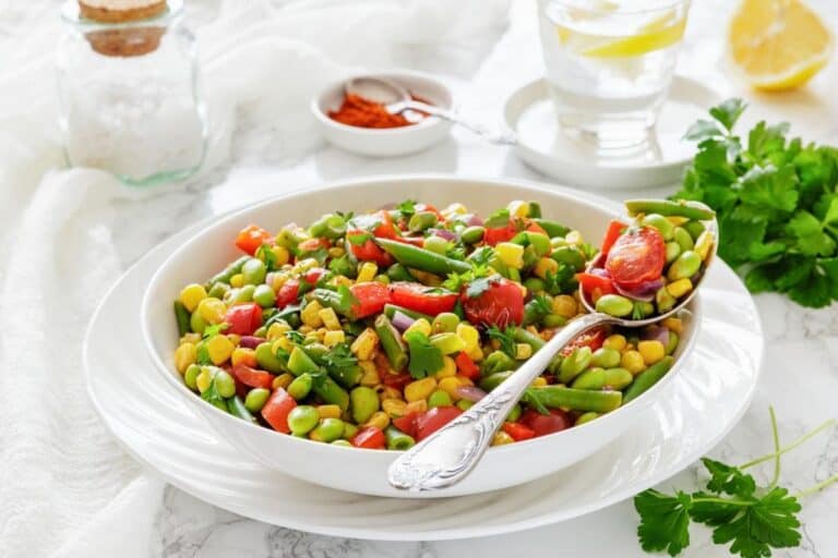 A white bowl filled with a colorful vegetable salad, including edamame, corn, cherry tomatoes, green beans, and red peppers, with a spoon resting inside.