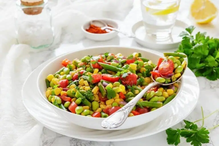 A white bowl filled with a colorful vegetable salad, including edamame, corn, cherry tomatoes, green beans, and red peppers, with a spoon resting inside.
