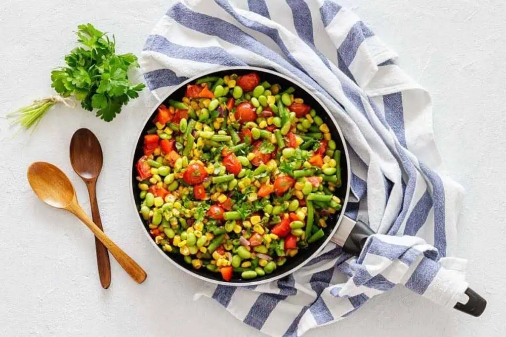 A skillet filled with a colorful vegetable stir-fry, including cherry tomatoes, edamame, corn, and peppers, sits on a white surface with a striped kitchen towel and two wooden spoons.