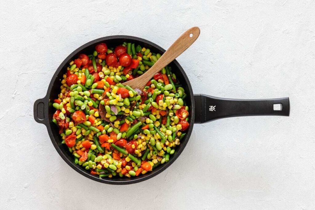 A black skillet filled with mixed vegetables, including corn, green beans, tomatoes, and edamame, with a wooden spoon on a light background.