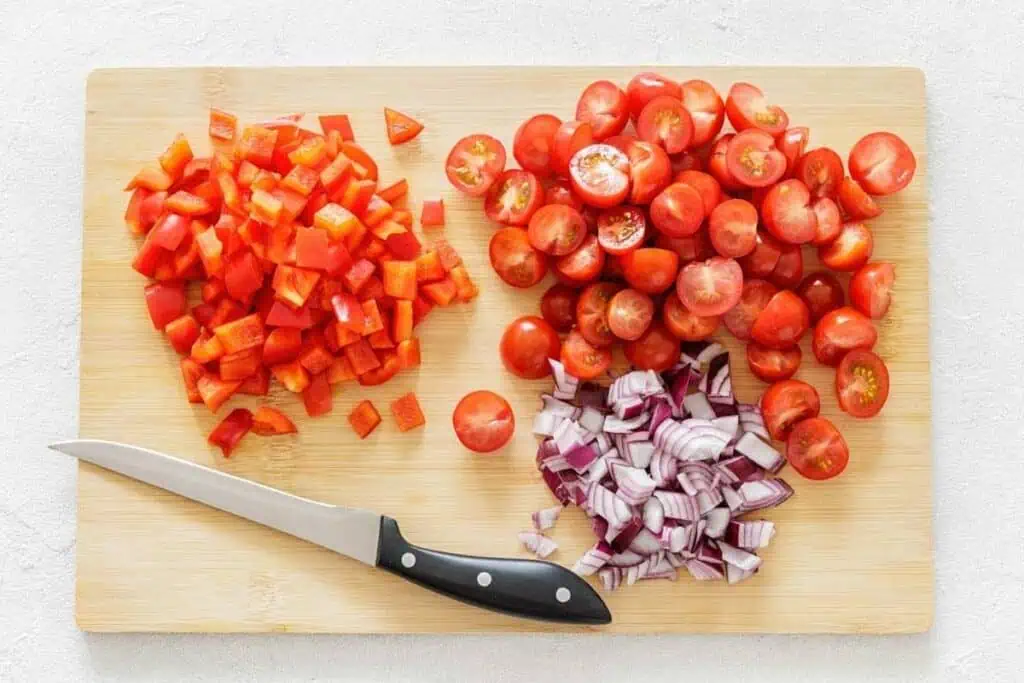 A cutting board with chopped red bell pepper, halved cherry tomatoes, diced red onion, and a kitchen knife.