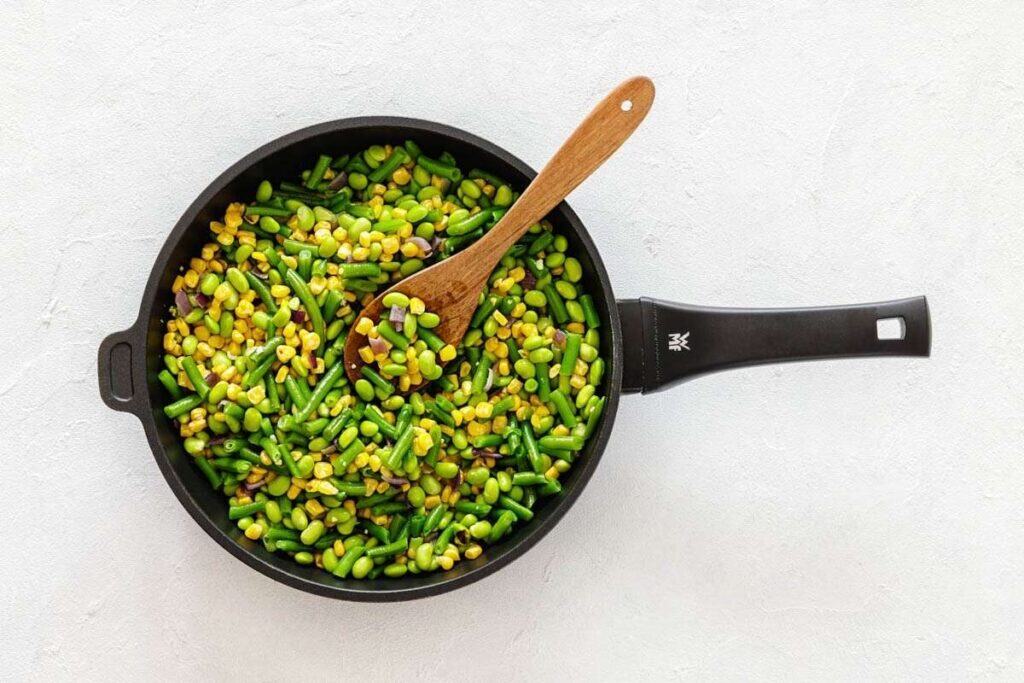 A black skillet filled with cooked mixed vegetables, including corn, green beans, and edamame, with a wooden spoon resting inside.