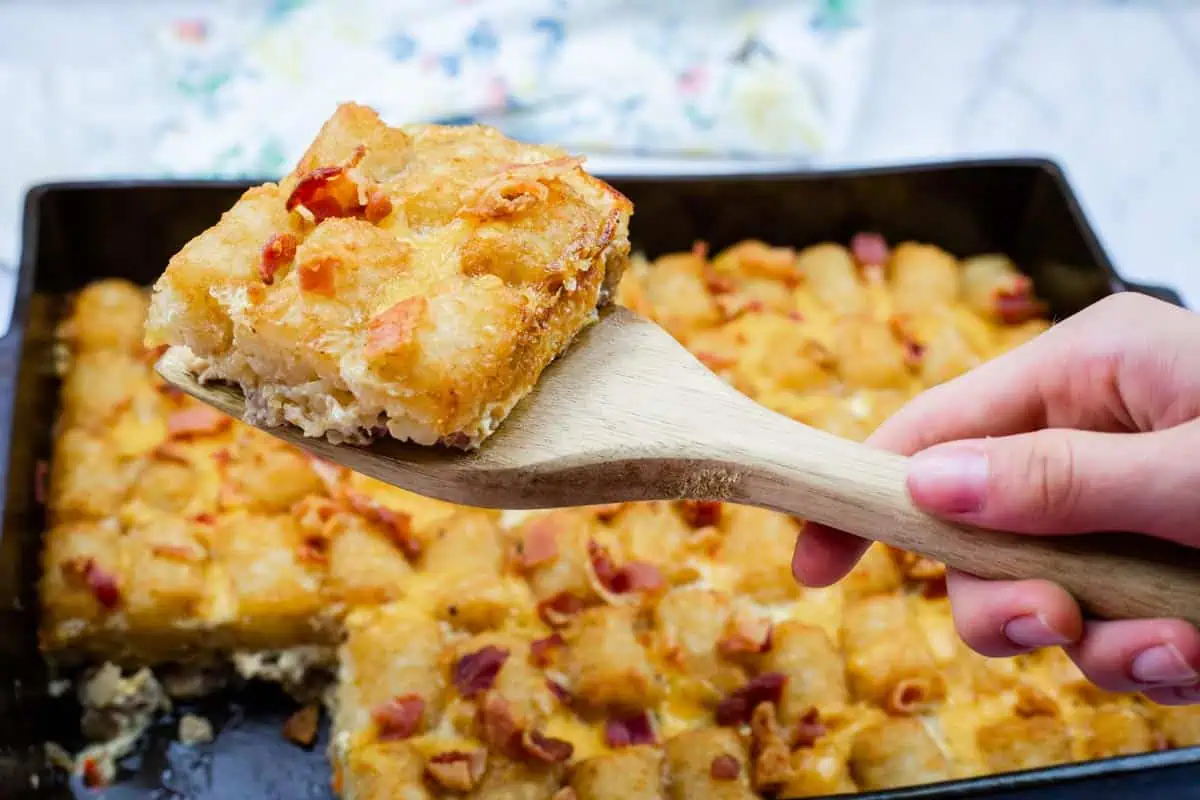 A hand holds a wooden spatula lifting a Cheesy Tater Tot Breakfast Casserole from a baking tray.