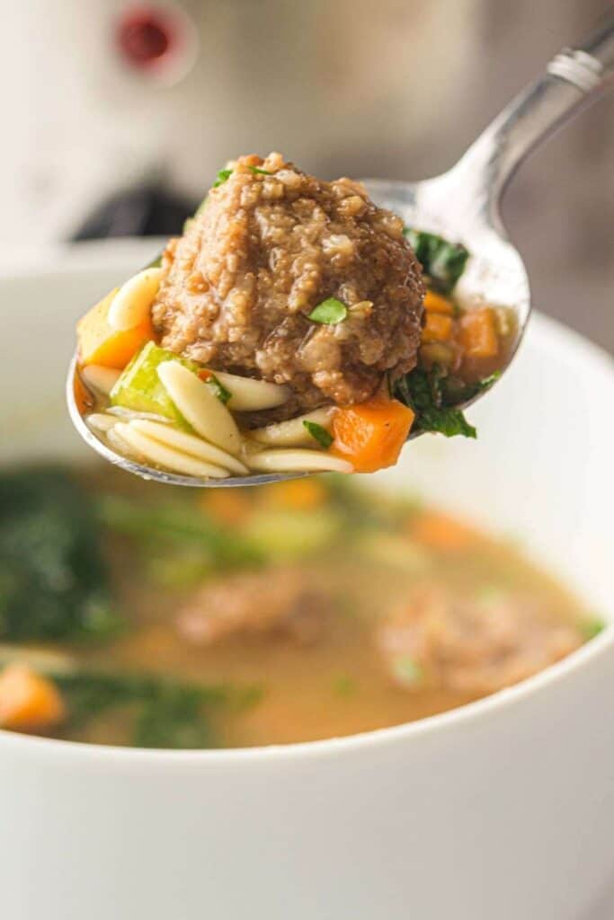 A close-up of a spoonful of soup with a meatball, pasta, carrots, celery, and greens held over a white bowl of soup.