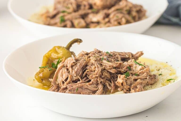 A white bowl filled with shredded beef, a whole pepper, and mashed potatoes, garnished with chopped herbs.