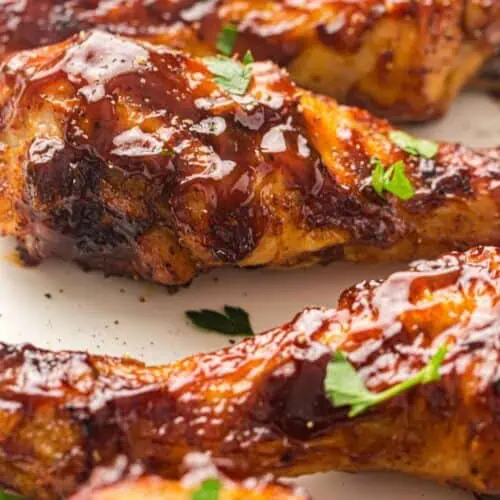 Close-up of baked chicken drumsticks coated in barbecue sauce and garnished with chopped parsley on a white plate.