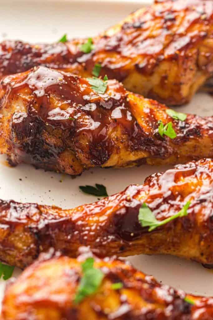 Close-up of baked chicken drumsticks coated in barbecue sauce and garnished with chopped parsley on a white plate.