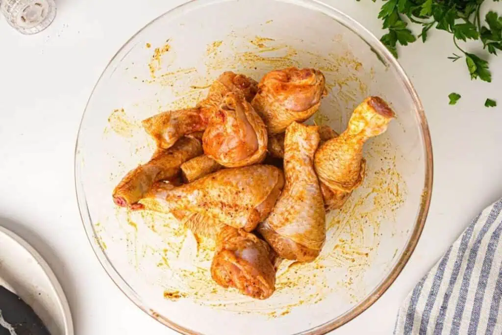 Raw chicken drumsticks coated in a seasoning marinade in a glass bowl, with chopped parsley and a striped cloth nearby on a white surface.