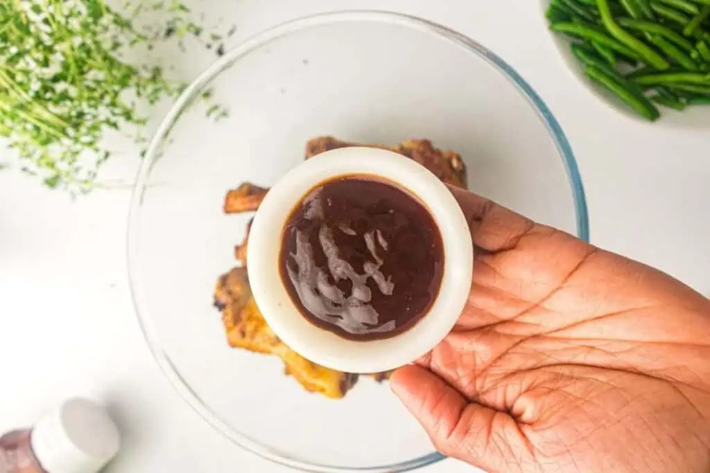 A hand holds a small white bowl of barbecue sauce above a glass bowl with cooked chicken pieces, with green beans and herbs visible nearby on a white surface.