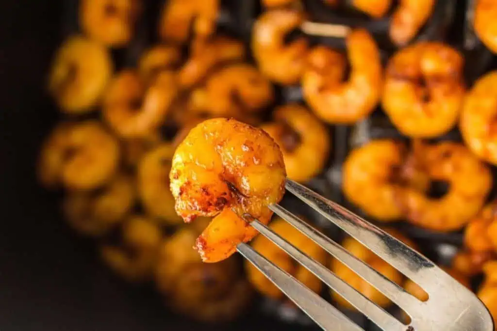 A close-up of a fork holding a seasoned, cooked shrimp with more shrimp in the blurred background.