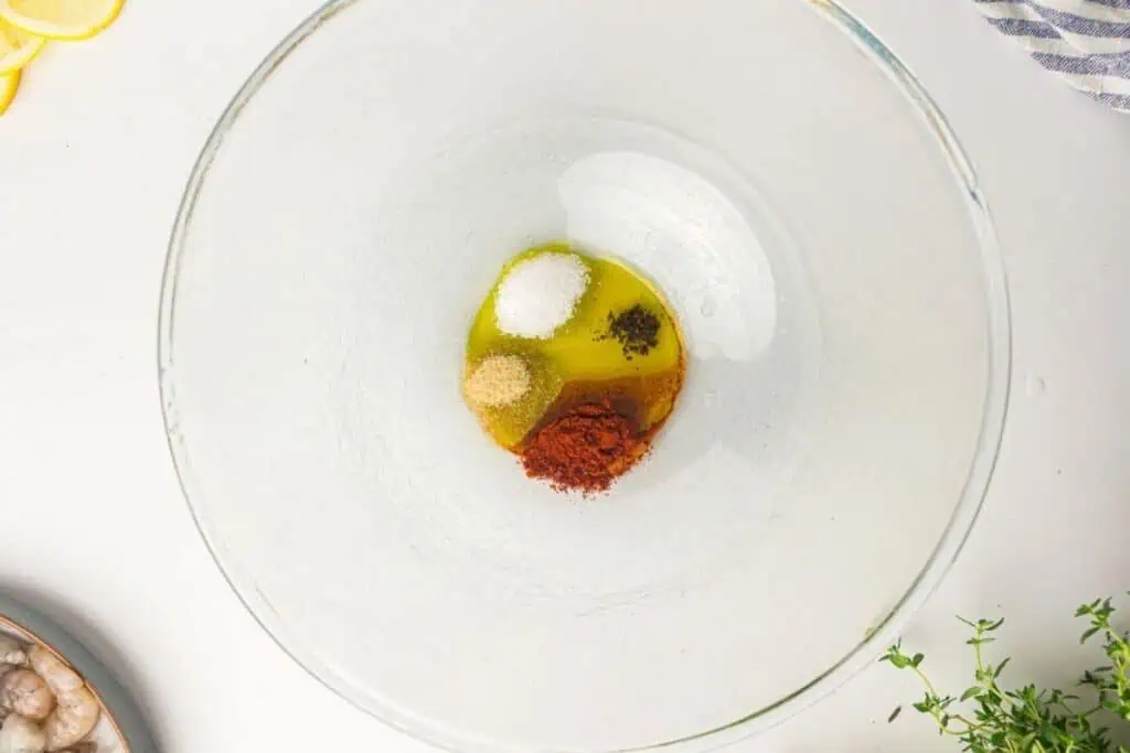 A glass mixing bowl containing olive oil, paprika, salt, black pepper, and garlic powder on a white surface.