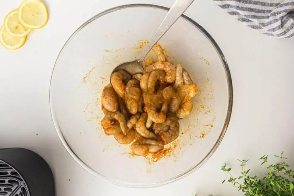 Raw shrimp coated with seasoning in a glass bowl, mixed with a metal spoon; lemon slices, herbs, and a striped cloth are nearby on a white surface.