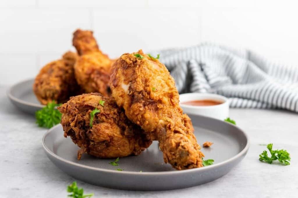 Two plates with pieces of fried chicken drumsticks garnished with parsley, accompanied by a small bowl of dipping sauce; striped cloth in the background.