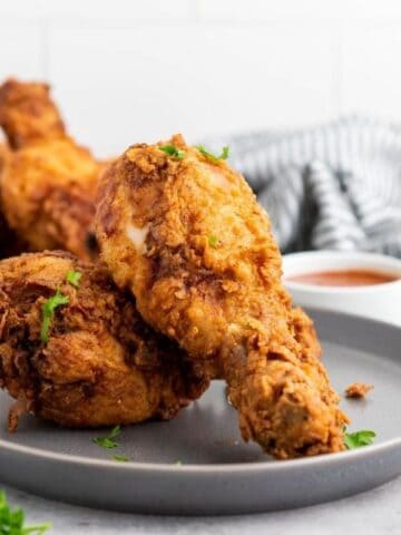 Two plates with pieces of fried chicken drumsticks garnished with parsley, accompanied by a small bowl of dipping sauce; striped cloth in the background.