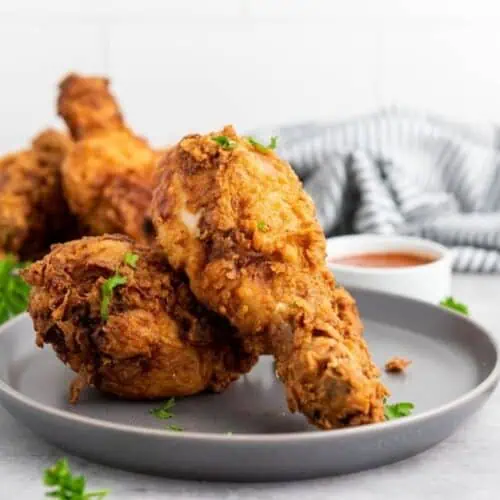 Two plates with pieces of fried chicken drumsticks garnished with parsley, accompanied by a small bowl of dipping sauce; striped cloth in the background.