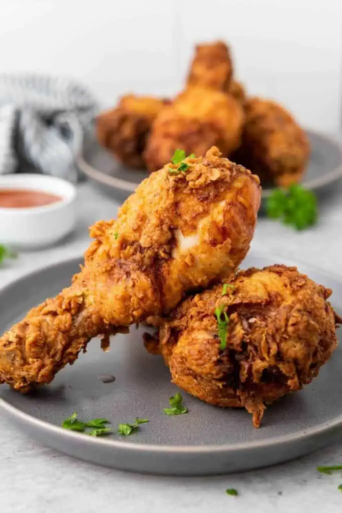 Two pieces of crispy fried chicken drumsticks on a gray plate, garnished with chopped herbs, with more chicken and sauce in the background.