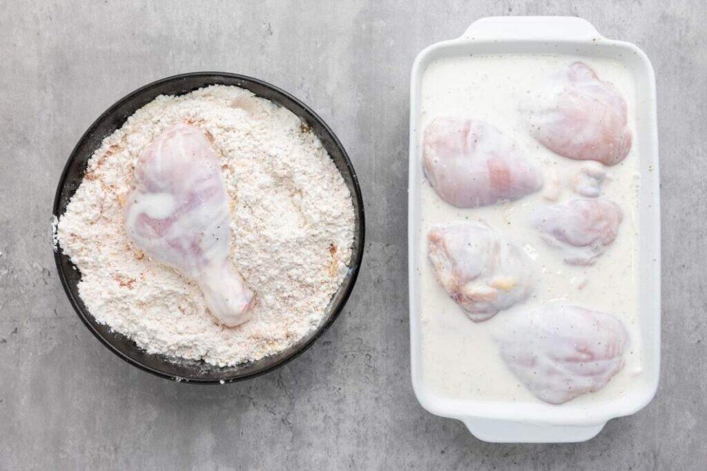 Raw chicken pieces in a white dish with marinade next to a bowl of flour coating with a chicken drumstick placed in the flour.
