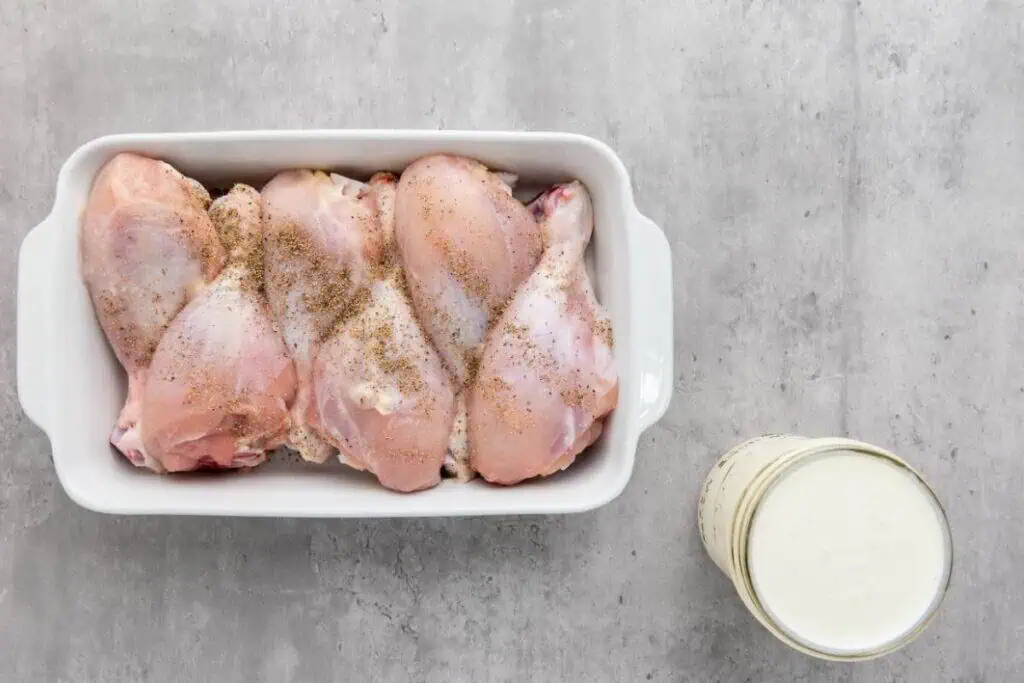 Four raw chicken drumsticks seasoned with pepper in a white baking dish, next to a glass jar filled with buttermilk, on a gray surface.