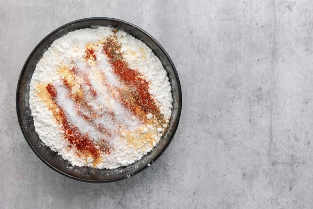 A black bowl filled with flour and various spices, including paprika, salt, and pepper, on a gray surface.