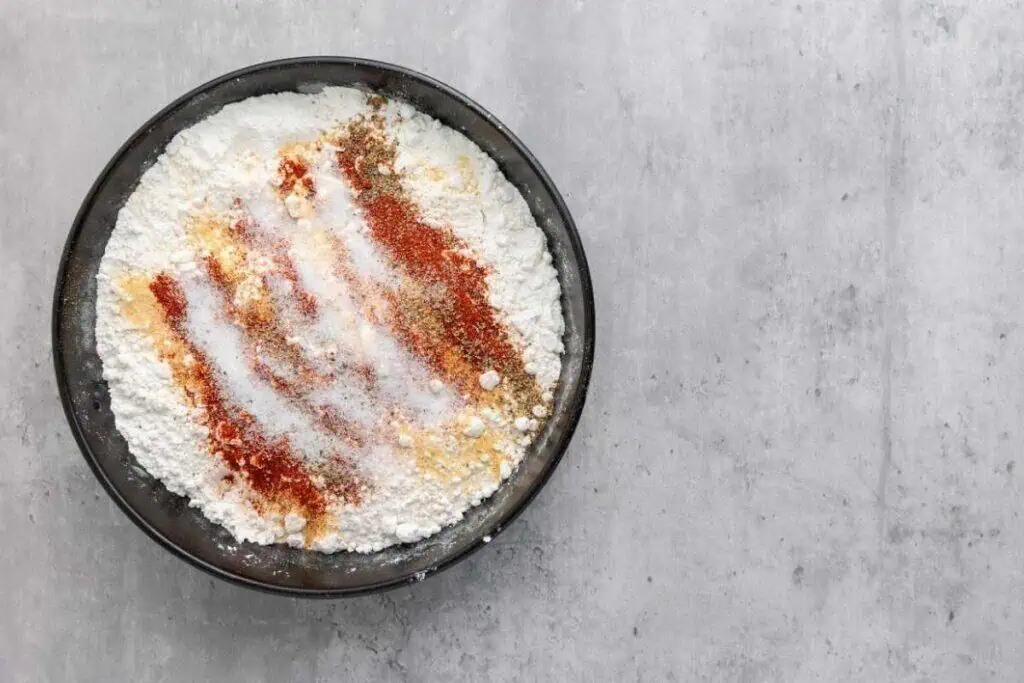 A black bowl filled with flour and various spices, including paprika, salt, and pepper, on a gray surface.