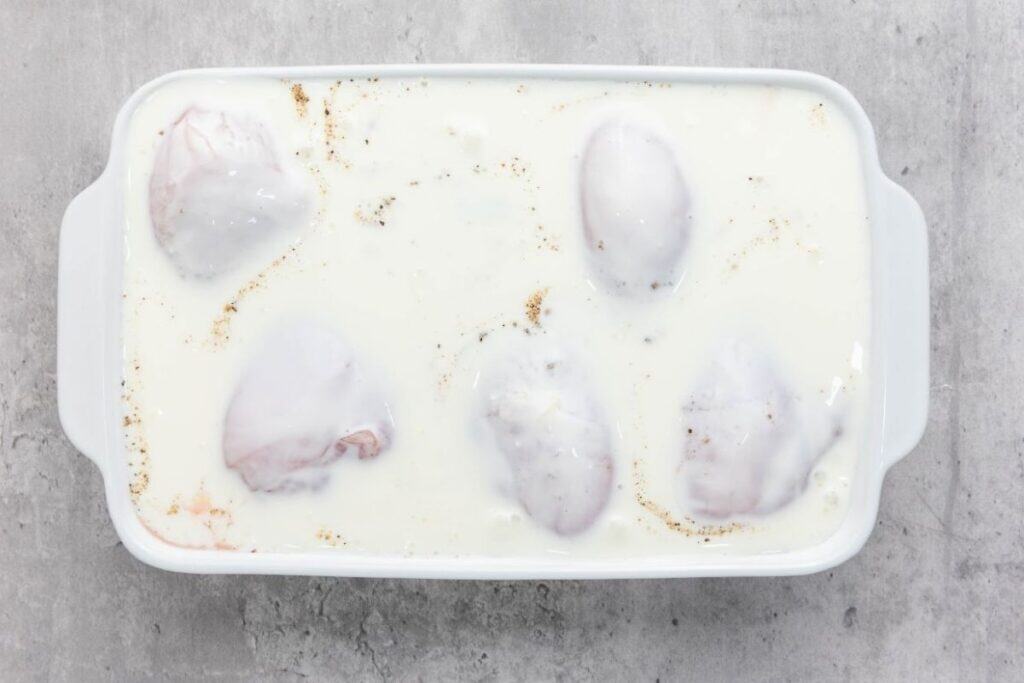 Five raw chicken breasts are covered in a white creamy liquid in a rectangular white baking dish on a gray surface.