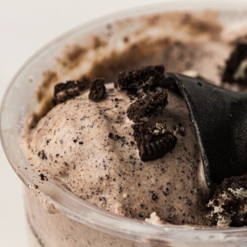 Close-up of cookies and cream ice cream in a cup, topped with crushed chocolate sandwich cookies and a black plastic spoon.
