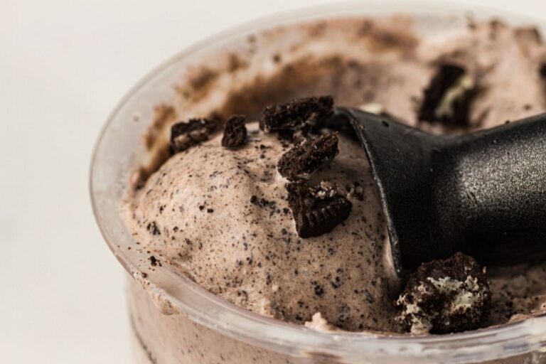Close-up of cookies and cream ice cream in a cup, topped with crushed chocolate sandwich cookies and a black plastic spoon.
