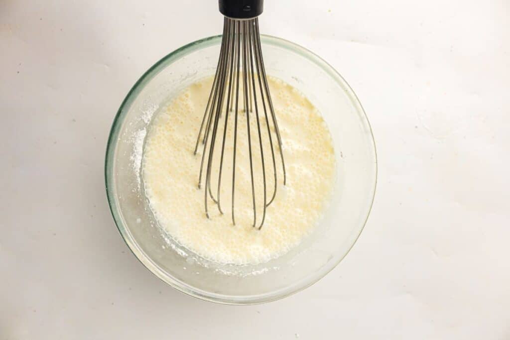 A metal whisk in a glass bowl containing a pale, creamy mixture on a white surface.