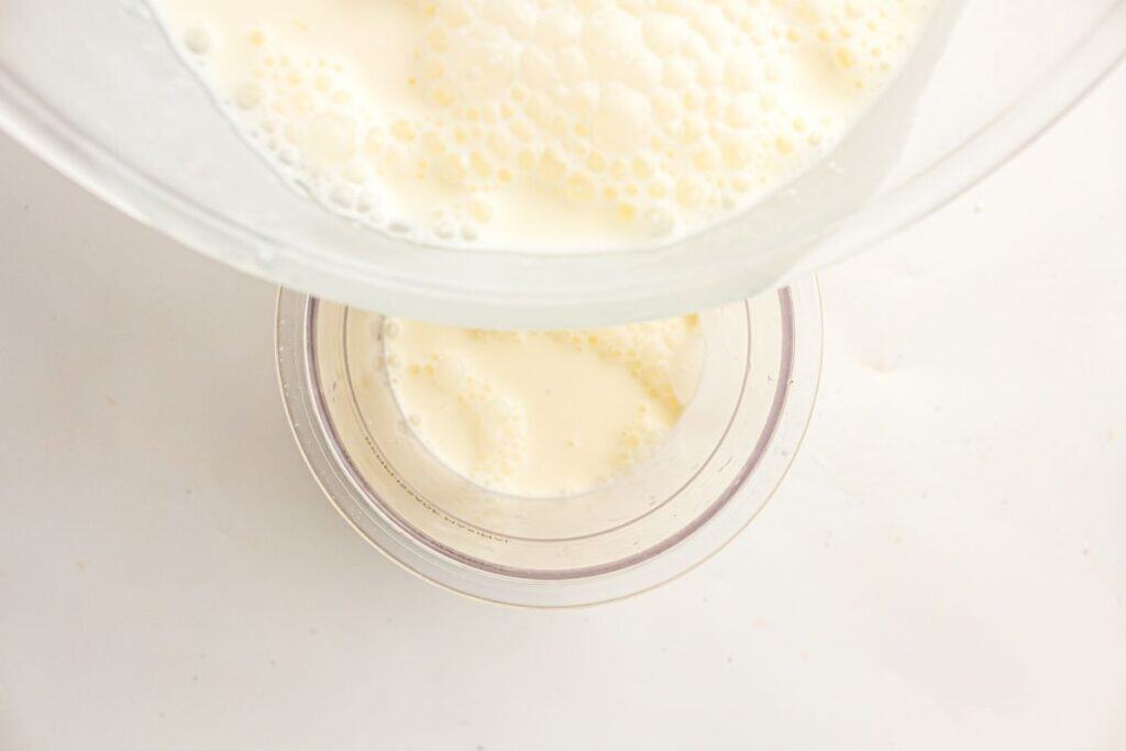 A glass bowl is pouring a creamy liquid, possibly milk or custard, into a smaller clear container on a white surface.