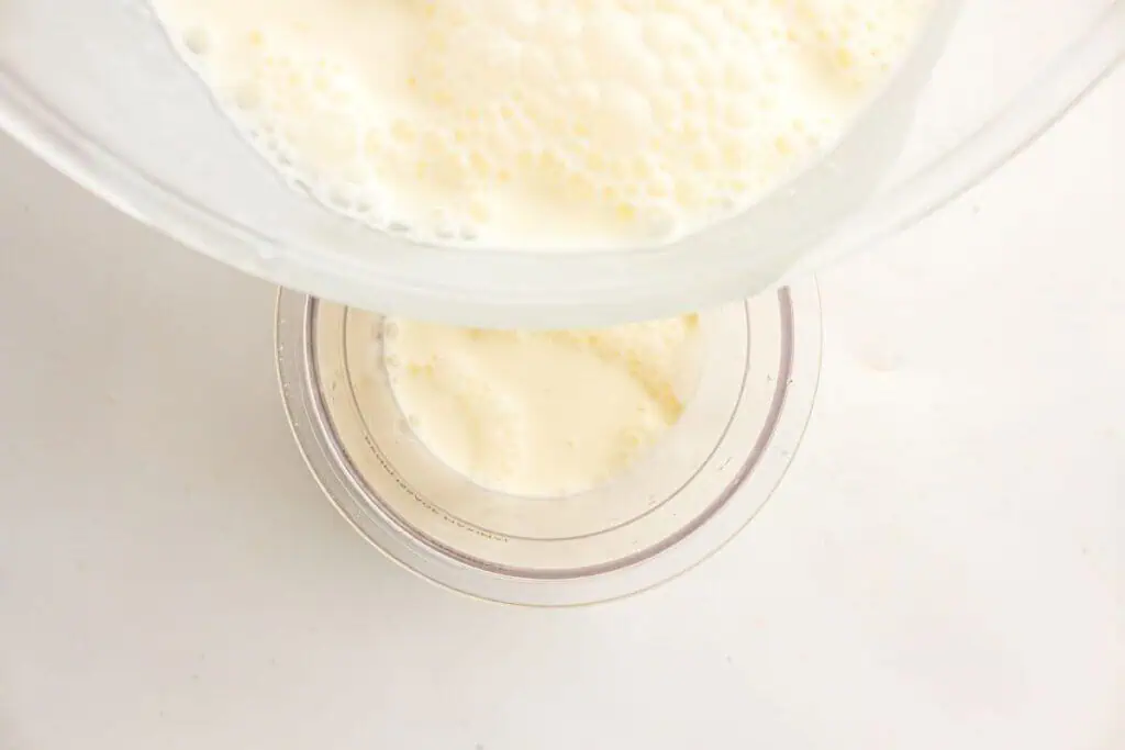 A glass bowl is pouring a creamy liquid, possibly milk or custard, into a smaller clear container on a white surface.