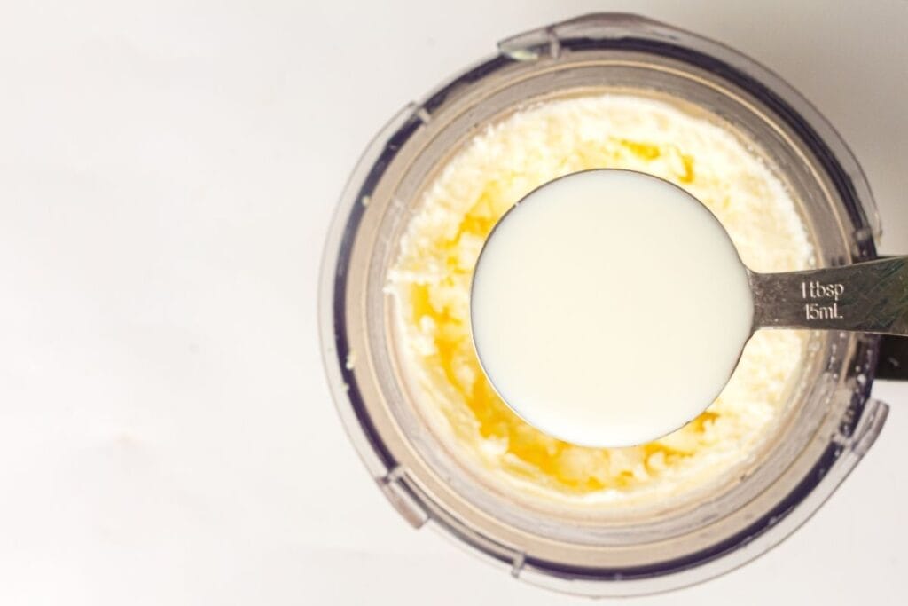 A metal tablespoon filled with milk held above a food processor containing a creamy mixture.