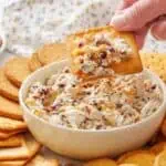 A hand dips a cracker into a bowl of cranberry cheese spread, surrounded by assorted crackers on a plate.