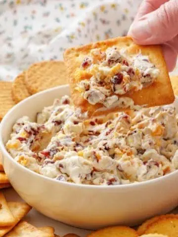 A hand dips a cracker into a bowl of cranberry cheese spread, surrounded by assorted crackers on a plate.