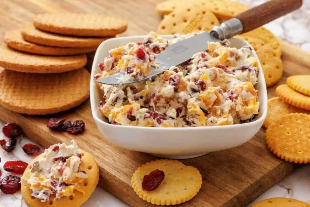 A square bowl of cheese and cranberry spread with a knife rests on a wooden board, surrounded by assorted round crackers and dried cranberries.