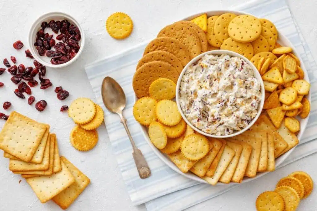 A plate of assorted crackers surrounds a bowl of creamy dip, with a spoon and a small bowl of dried cranberries on a striped napkin. Crackers and cranberries are scattered nearby.