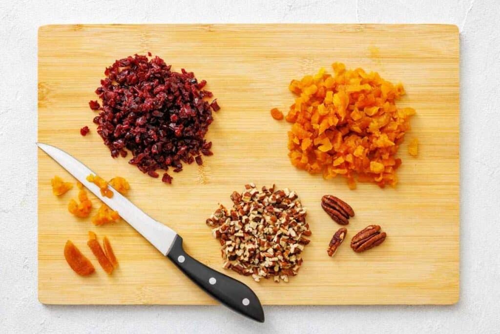 A cutting board with chopped dried cranberries, dried apricots, and pecans, along with a knife and unchopped apricots and pecans.