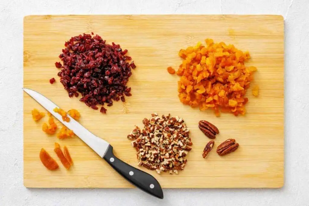 A cutting board with chopped dried cranberries, dried apricots, and pecans, along with a knife and unchopped apricots and pecans.