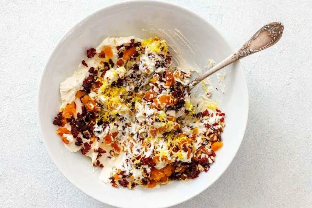 A white bowl containing cream cheese, chopped dried fruit, lemon zest, and nuts, with a silver spoon resting inside.