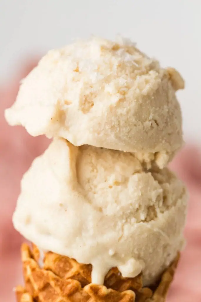 Two scoops of vanilla ice cream stacked on a waffle cone, with a soft focus pink background.
