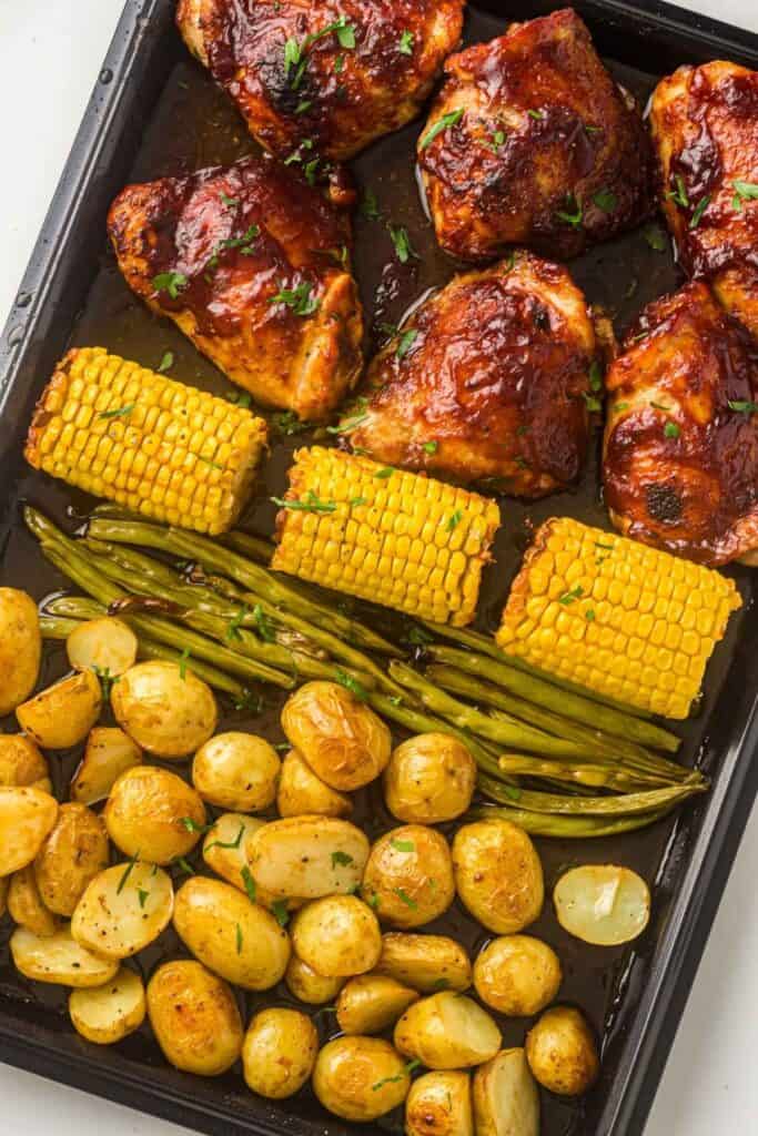 Sheet pan with roasted BBQ chicken thighs, corn on the cob, green beans, and seasoned baby potatoes.