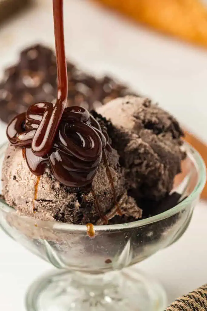 Two scoops of cookies and cream ice cream in a glass bowl with chocolate syrup being poured on top.